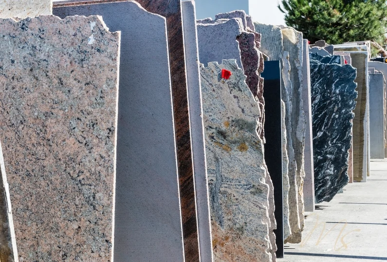 Outdoor display of various granite slabs standing upright in a row. The slabs showcase a range of colours and patterns, including shades of grey, black, and brown. The scene is set in a storage or sales yard with trees visible in the background.