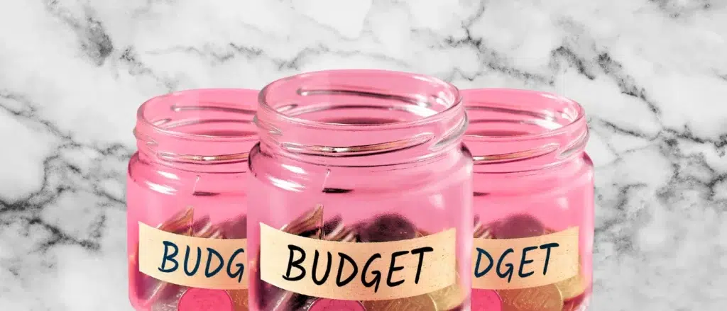 Three pink glass jars labeled "BUDGET," partially filled with coins, set against a marble background.