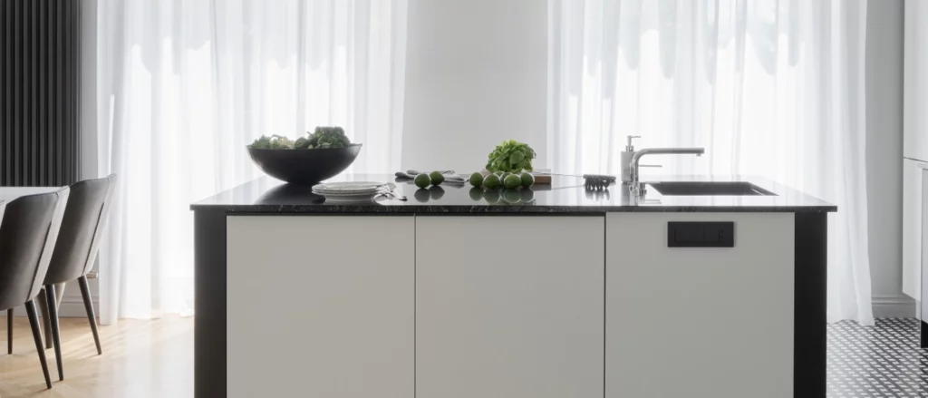  A modern kitchen island with a black countertop and light cabinetry. The counter holds limes, fresh herbs, a bowl of vegetables, plates, and cutlery. A stainless steel sink is integrated, with soft light filtering through white curtains in the background.