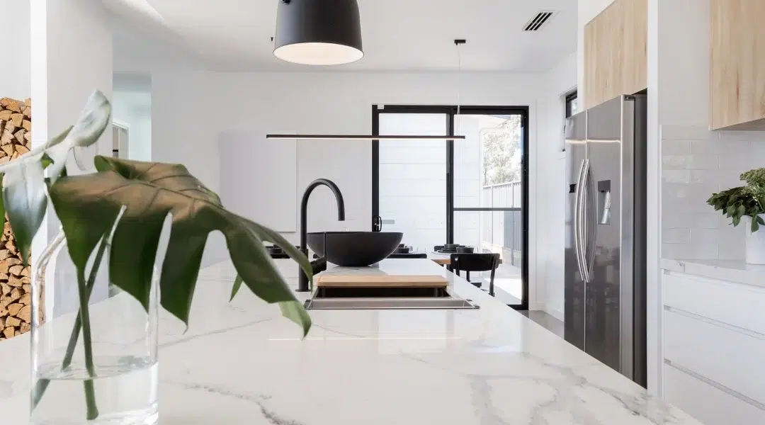 A modern kitchen with a sleek marble countertop, black fixtures, and wood-accented cabinets. In the foreground, a glass vase holds large green leaves. The background features a stainless steel refrigerator, white tile backsplash, and minimalistic decor.