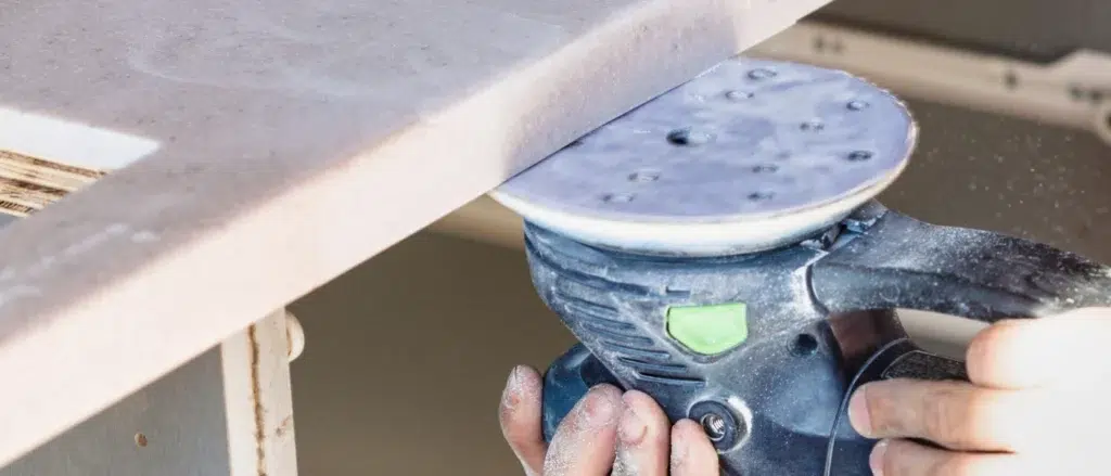 Close-up of a person using a sander on the edge of a kitchen countertop, creating dust.